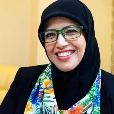 A woman wearing glasses, a black headscarf and a floral scarf smiles, photographed against a yellow background.
