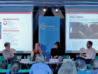 Panel discussion with four people on stage in front of an "elbarlament cultures of democracy" banner, with screens showing the headline "Culture matters!" and an audience in the foreground.