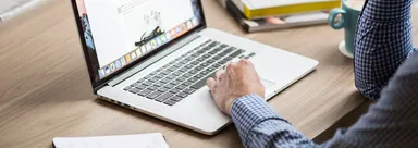 A hand rests on a laptop trackpad beside a keyboard, notebook and cup on a wooden desk.