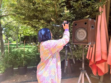 A woman in a headscarf holds up a camera on a monopod near a large speaker mounted on a tripod, in a leafy garden.