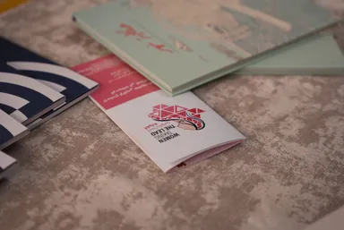 A stack of booklets on a textured surface, the top one showing a logo of hands and colorful triangles above the text "WOMEN TAKING THE LEAD", appearing upside down, beside striped and pastel booklets.