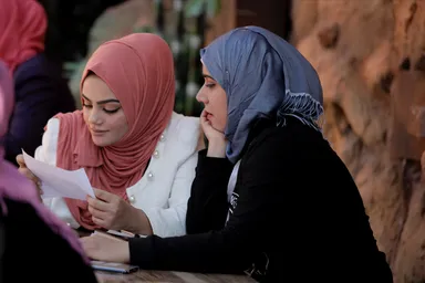 Two women sit at a table outdoors, one holding and reading a sheet of paper while the other rests her chin on her hand, in front of a rocky wall.