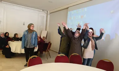 A group of people stand raising joined hands in a room, with seated attendees and a screen showing a desktop display behind them.