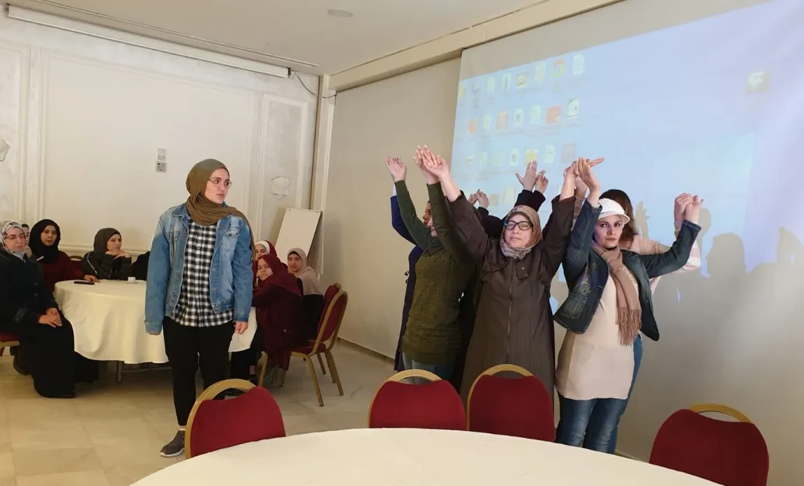A group of people stand raising joined hands in a room, with seated attendees and a screen showing a desktop display behind them.