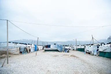Rows of tarpaulin-covered shelters in a camp, with water tanks and cables between them and snow-capped mountains in the distance.