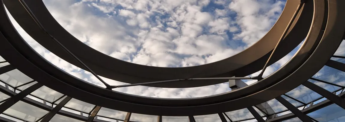 View upward through the steel and glass dome of a parliament building at a cloudy blue sky, showing the curved ramp structure and mirrored cladding.