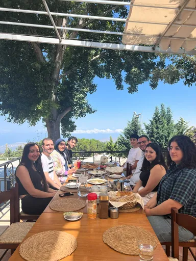 A group of people sit at a long outdoor dining table under trees with a sea view, plates of food and drinks in front of them.
