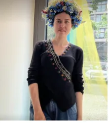 A person wears a floral crown of blue and white flowers over a black top with an embroidered sash, standing in front of a window.