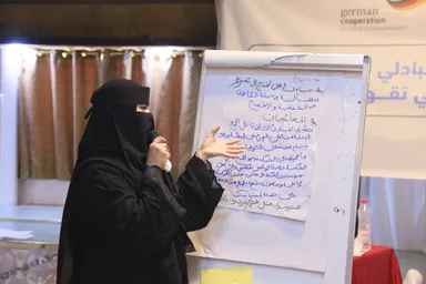 A woman in a face-covering veil holds a microphone and gestures toward a flipchart with handwritten Arabic text, beside a banner reading "german cooperation".
