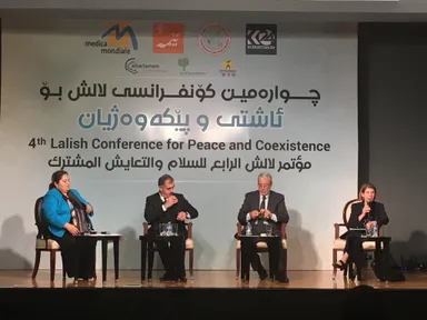 Four people seated on a stage in front of a banner with several logos and the text "4th Lalish Conference for Peace and Coexistence" in English, Arabic and Kurdish.