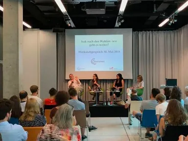 Panel of four people seated on a stage in front of an audience, with a projected slide reading "Irak nach den Wahlen - wie geht es weiter?" and "Werkstattgespräch 30. Mai 2018".