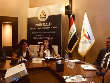 Panel discussion at a conference table with three people and microphones, in front of a banner reading "2019 R.C.D SECURITY AND ECONOMIC FORUM, BAGHDAD - BABYLON ROTANA HOTEL, IRAQ IS RECOVERING", flanked by the Iraqi flag and a dialogue-centre flag.
