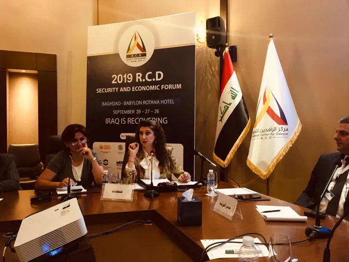 Panel discussion at a conference table with three people and microphones, in front of a banner reading "2019 R.C.D SECURITY AND ECONOMIC FORUM, BAGHDAD - BABYLON ROTANA HOTEL, IRAQ IS RECOVERING", flanked by the Iraqi flag and a dialogue-centre flag.