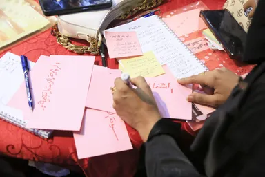 Hands write on pink sticky notes with Arabic text at a table covered with notebooks, a phone and a handbag.