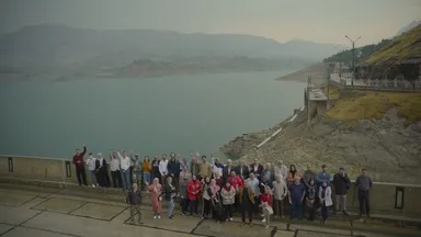 A large group of people stand along a lakeside walkway, some waving, with hills and a dam structure in the background.