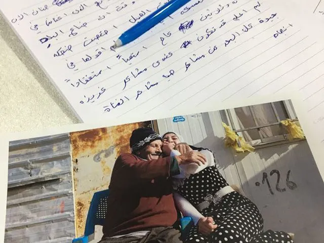 Handwritten Arabic notes and a pen rest on a page above a printed photo of an older man embracing a young woman outside a shelter marked "0.126".