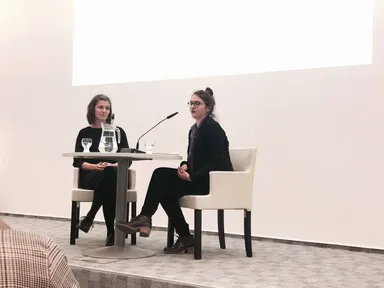 Two women seated at a small table on a stage, one speaking into a microphone.