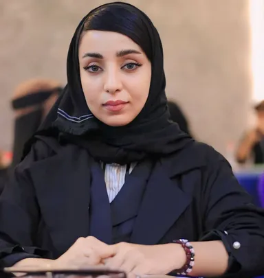 A woman in a black headscarf and blazer sits with hands folded on a table, with blurred figures visible in the background.