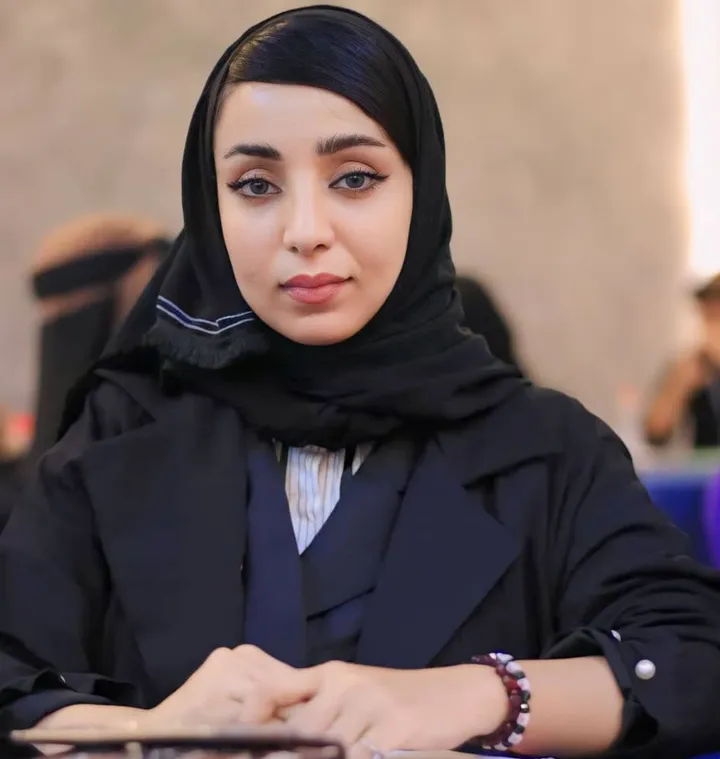 A woman in a black headscarf and blazer sits with hands folded on a table, with blurred figures visible in the background.