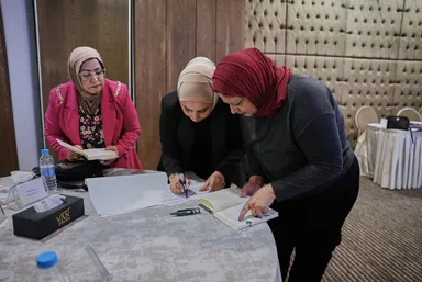 Three women in headscarves lean over a table, one writing in an open book while another looks on.