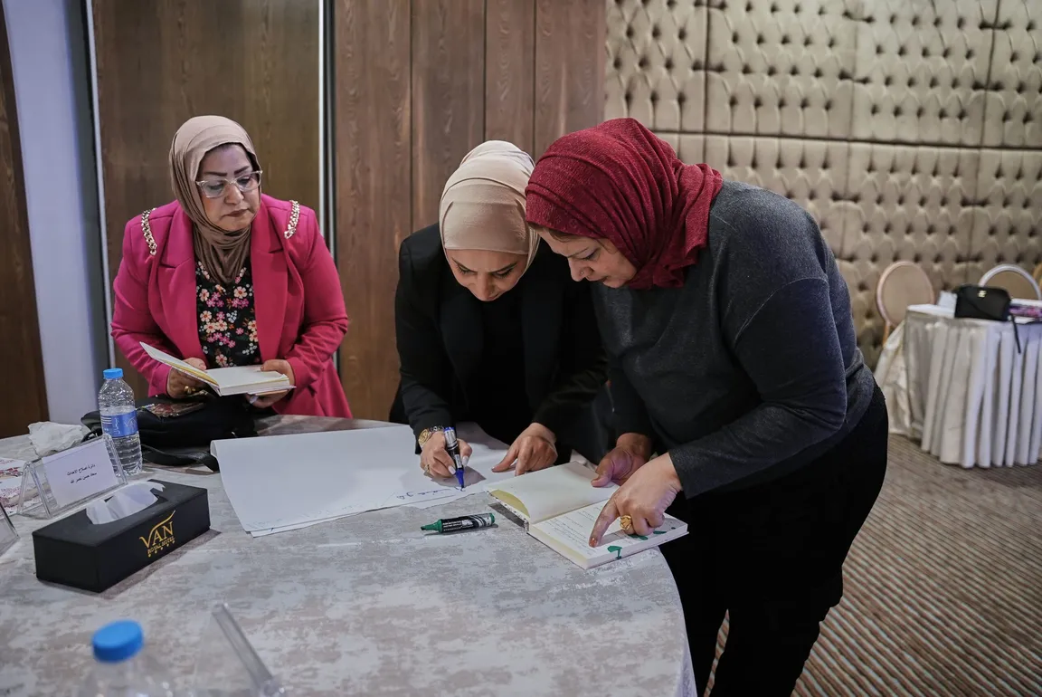 Three women in headscarves lean over a table, one writing in an open book while another looks on.