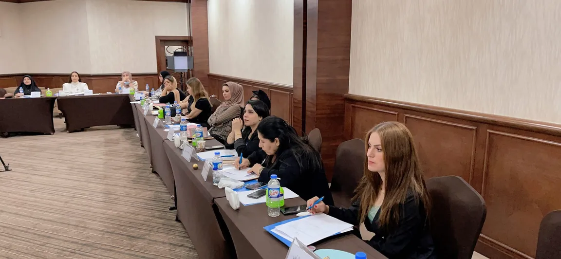 Several people sit along a long conference table with papers, water bottles and name cards, in a meeting room.