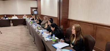 Several people sit along a long conference table with papers, water bottles and name cards, in a meeting room.