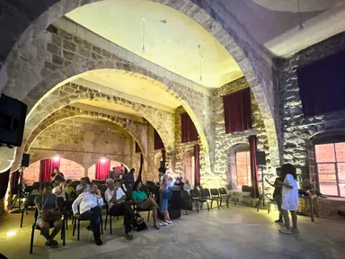 Interior of a vaulted stone hall with red curtains and rows of chairs, with two people standing near a laptop and speaker on the right.