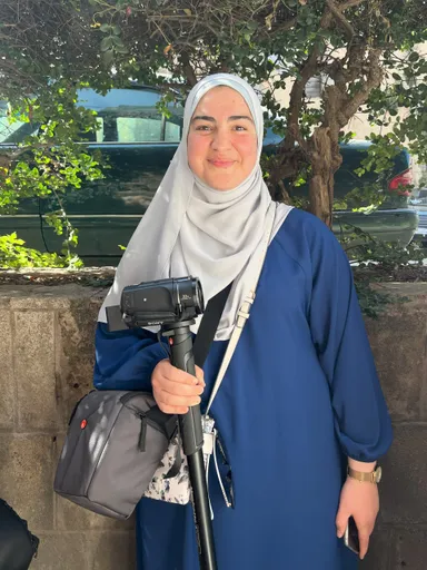 A smiling woman in a headscarf holds a video camera mounted on a handle, standing outdoors in front of a tree and a parked car.