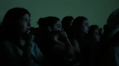 A row of people watching something intently in a dimly lit, teal-toned room.