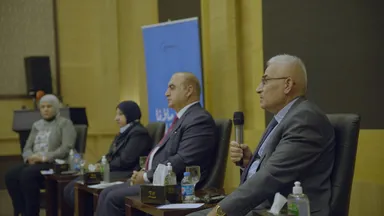 Four panelists sit at a table, a man speaking into a microphone, with water bottles and a tissue box on the table.
