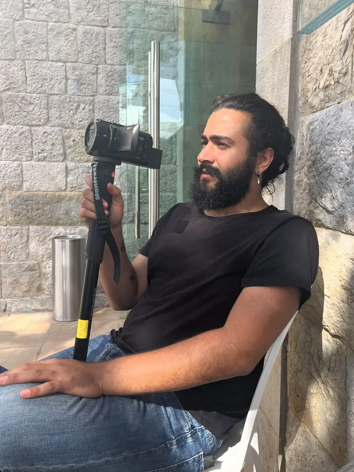 A man sits outdoors holding a video camera mounted on a monopod against a stone wall.