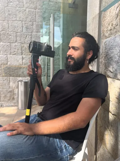 A man sits outdoors holding a video camera mounted on a monopod against a stone wall.