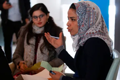 A woman wearing a patterned headscarf gestures while speaking, holding a pen and handwritten notes, as another woman listens.