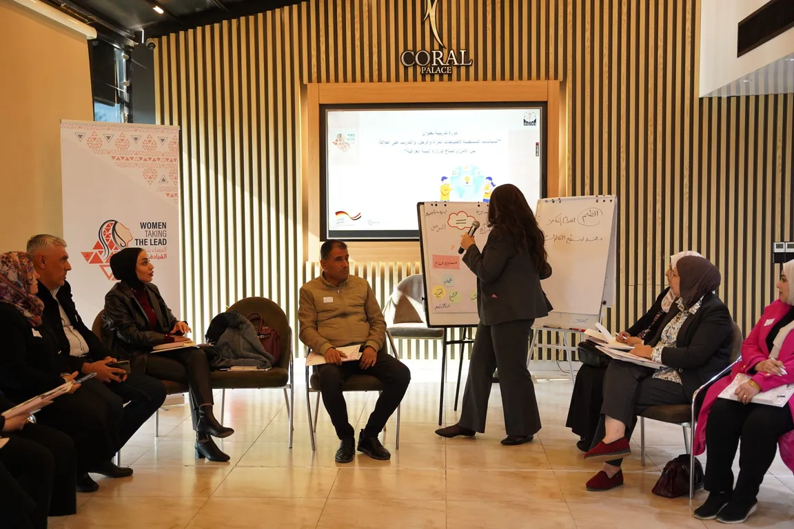 People sit in a conference room beneath a "Women Taking the Lead" banner while a woman stands writing on a flip chart in front of a screen with Arabic text.