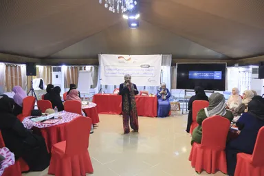A person speaks into a microphone in a hall where a group of people in headscarves sit at round tables facing a banner and a screen with Arabic text.