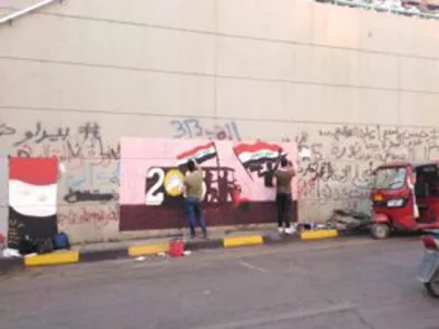 Two people paint a red-white-black flag onto a mural on a graffiti-covered wall, with a rickshaw parked on the street beside them.