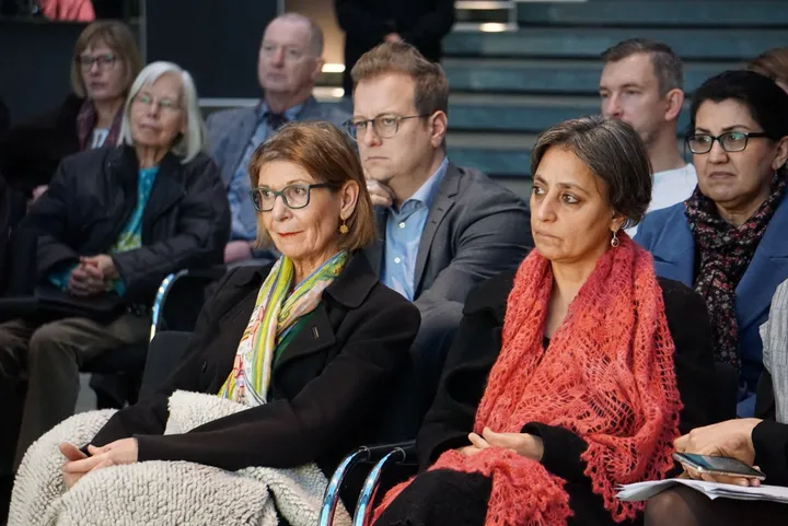 A row of seated people listening attentively during an indoor event, with stairs visible in the background.
