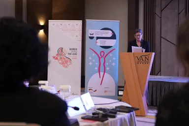 A woman speaks at a wooden lectern reading "VAN ROYAL HOTEL", beside two banners reading "WOMEN TAKING THE LEAD" and "Strengthening Participation, Peaceful Coexistence and Equality in Iraq", with two blurred heads in the foreground.