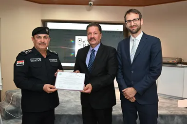 Two men in suits presenting a certificate to a police officer, with a screen showing a video-call interface in the background.