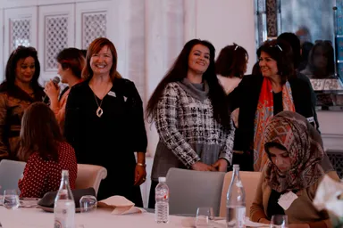 A group of people stand and sit around a long table set with water bottles and glasses, several smiling as one speaks into a microphone.