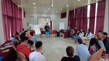 People seated in a circle in a room with a man standing near a screen and flip chart during a presentation.