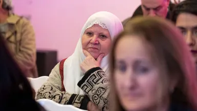 An older woman wearing a white headscarf rests her chin on her hand while seated among an audience.