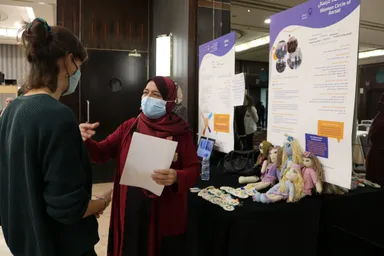 Two women wearing face masks talk at an event booth beside a poster reading "Women Circle of Aarsal", with knitted dolls and papers on the table.