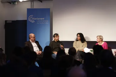 Four people sit on a panel stage in front of a blank projection screen and a banner reading "elbarlament cultures of democracy" with Arabic text, an audience seated in the foreground.