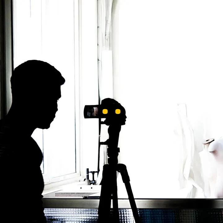Silhouette of a person next to a camera on a tripod, backlit by a bright window.