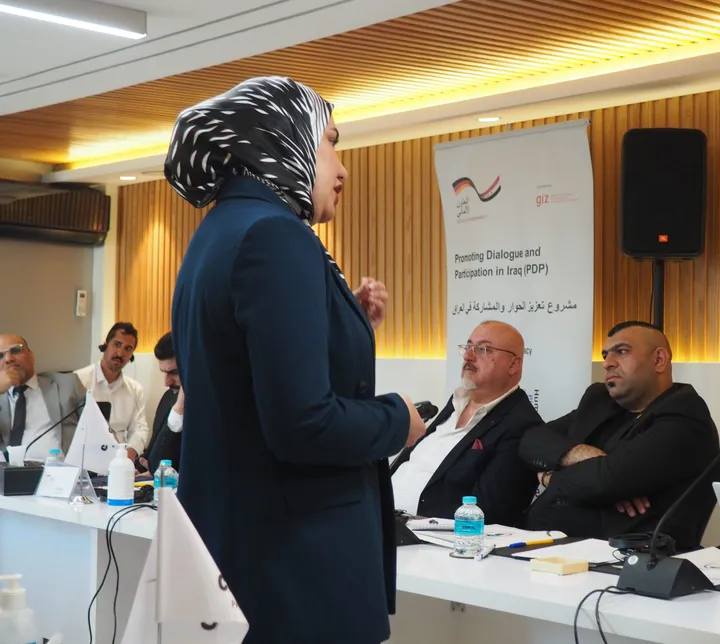A woman wearing a headscarf speaks while holding a microphone, with seated people and a banner reading "Promoting Dialogue and Participation in Iraq (PDP)" behind her.