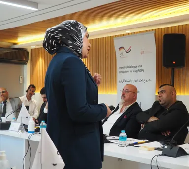 A woman wearing a headscarf speaks while holding a microphone, with seated people and a banner reading "Promoting Dialogue and Participation in Iraq (PDP)" behind her.