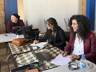 Three women sit at tables laughing and writing notes, with a laptop, notebooks and camera equipment in front of them.