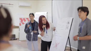 A woman speaking into a microphone beside a flip chart with hand-drawn flowers and notes, with two others standing nearby.